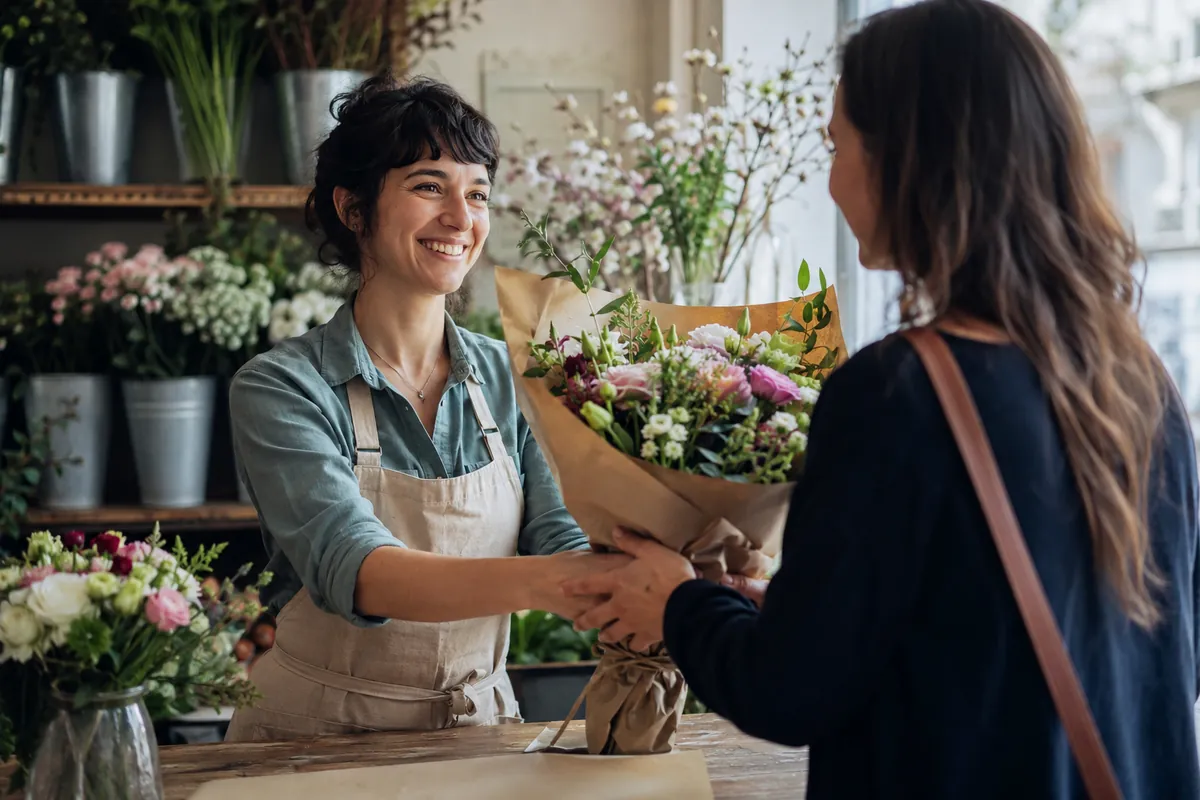 A local florist hands a wrapped seasonal bouquet across the counter to a customer