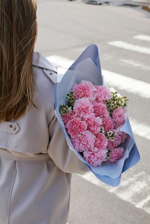 A woman in a beige coat carrying a wrapped bouquet of pink carnations